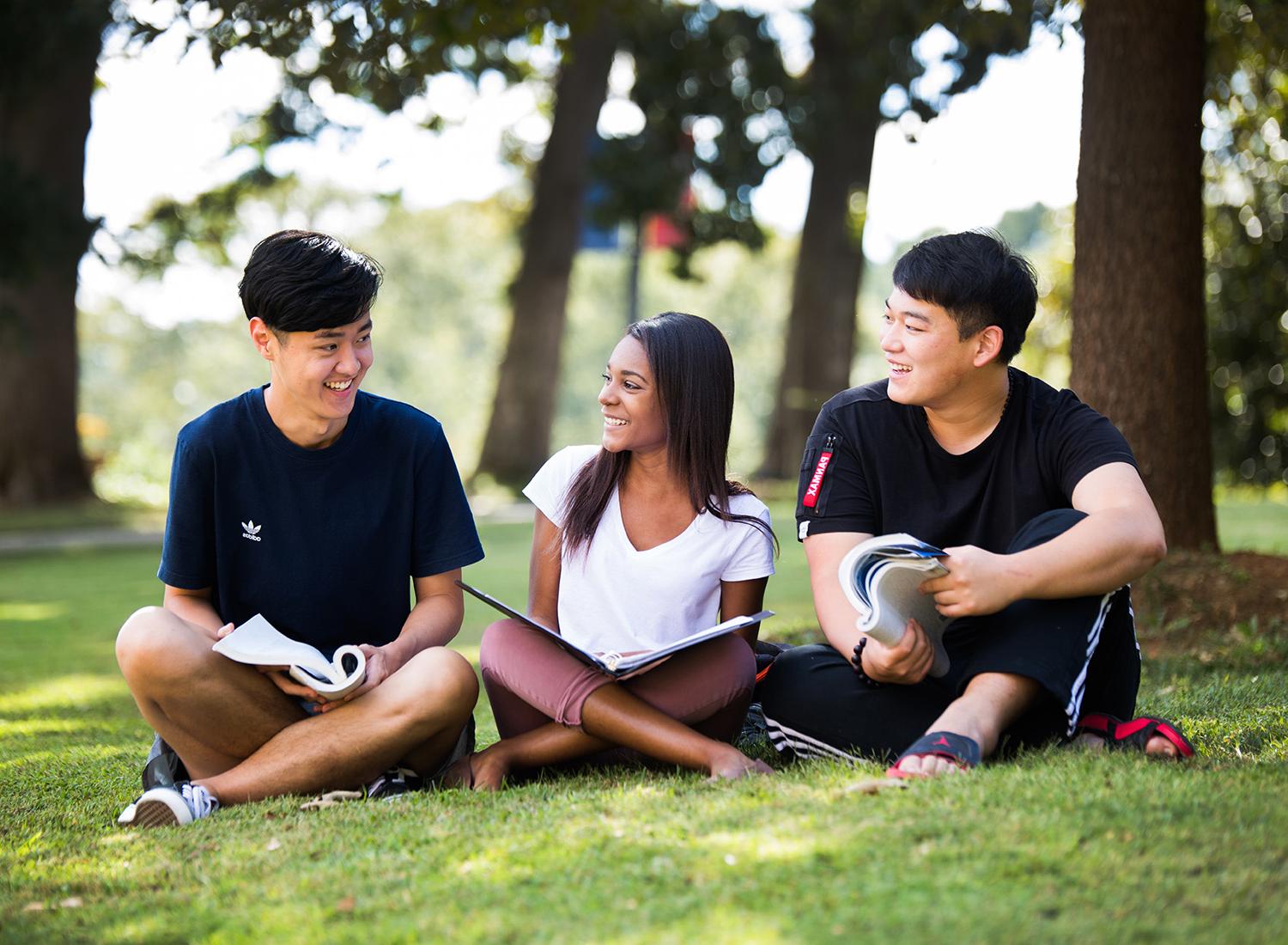 三个学生拿着课本坐在外面的草地上.